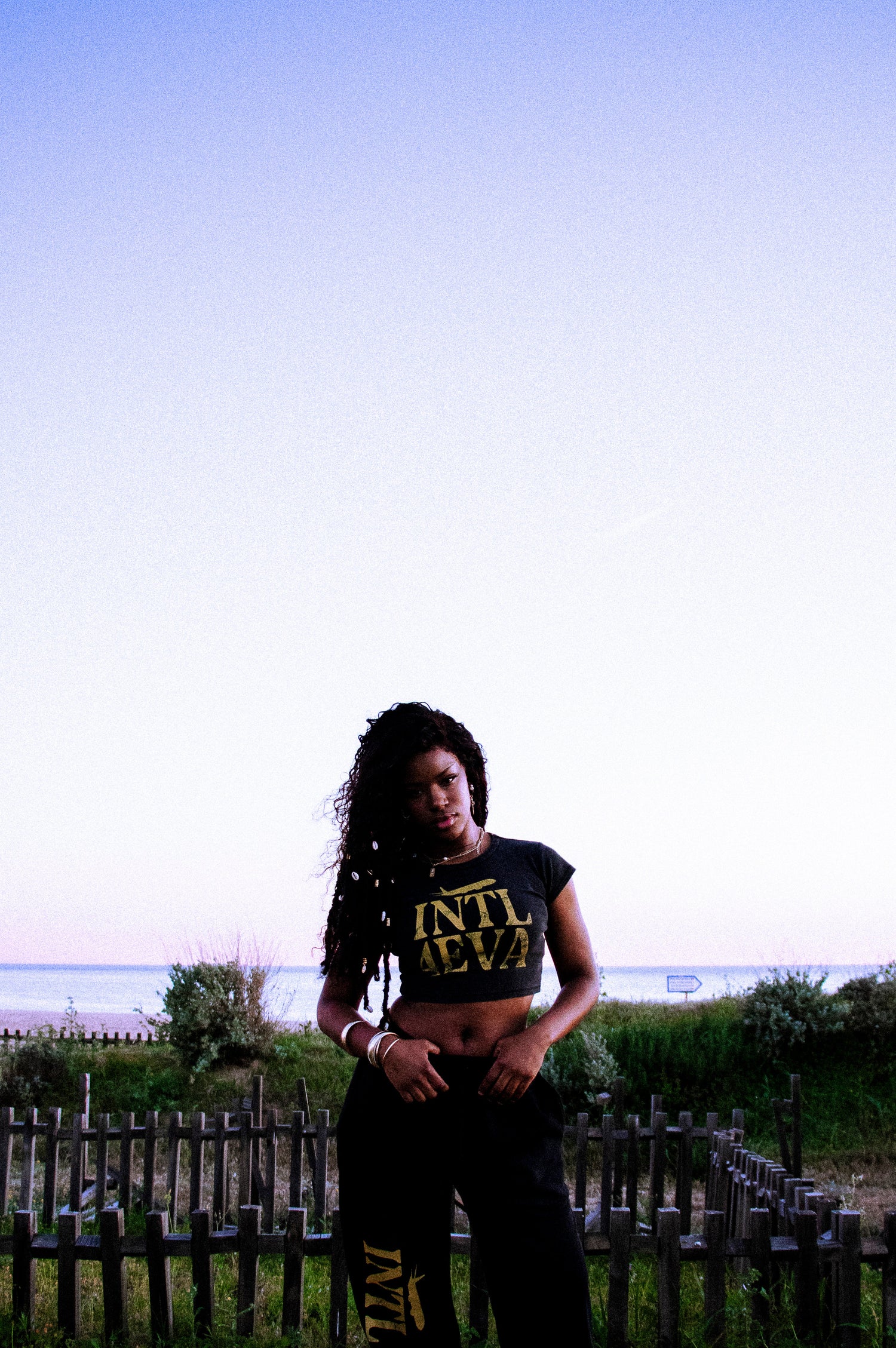 Person wearing a black outfit with text standing outdoors near a fence and grass.