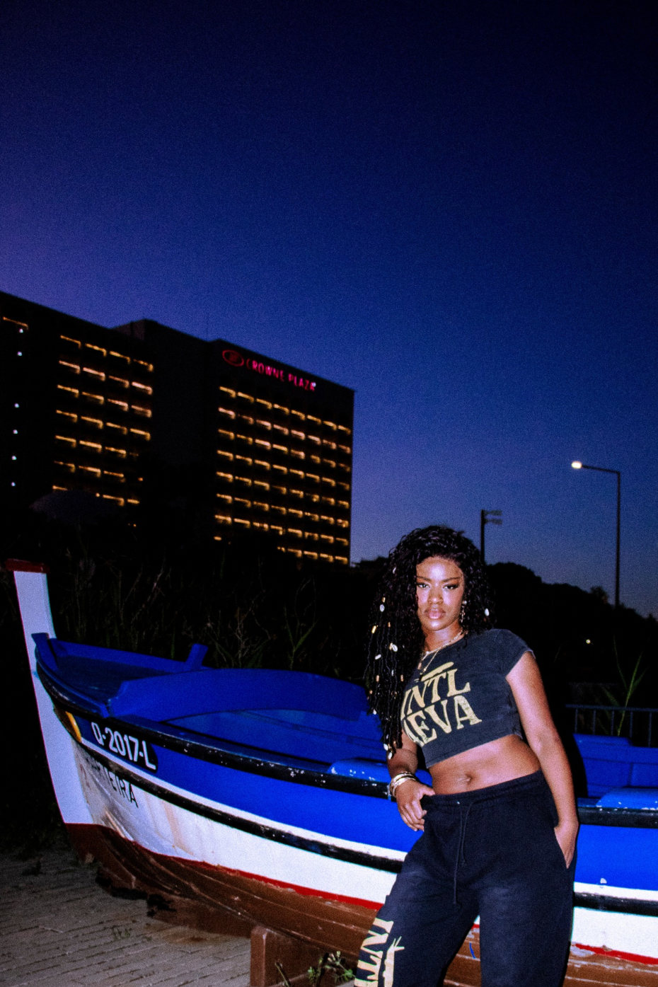 Woman standing next to a boat at night with buildings in the background with baby tee on 