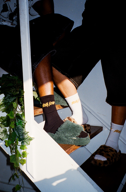 A pair of black ankle-length NEVA STOP socks with the brand's logo on the sole, worn by a person standing on a wooden staircase with plants in the background.