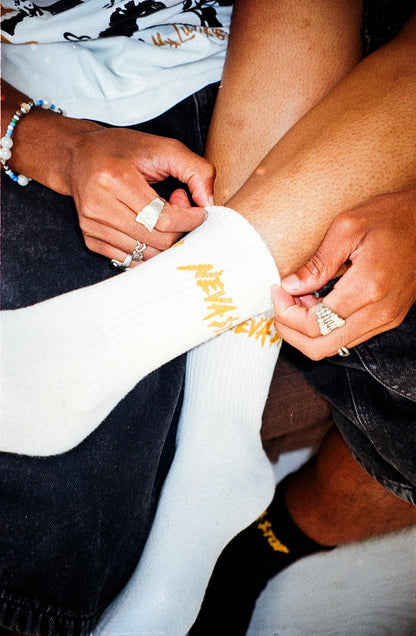 A person is wearing a white ankle-length sock with the text 'NEVA STOP' printed on it. The sock is partially rolled up, and a ring is visible on one of the fingers adjusting the sock.